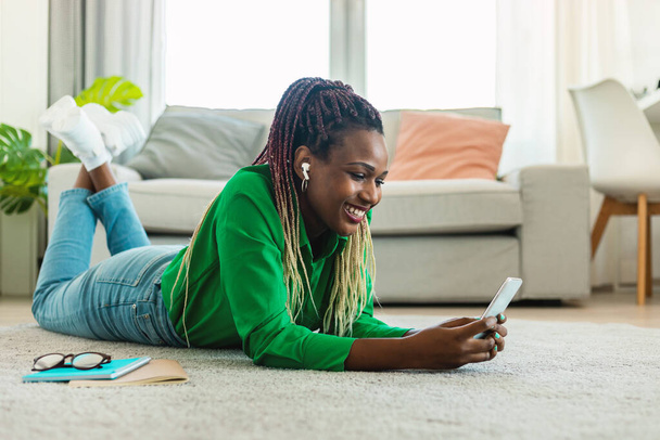 Excited african american lady in earphones using cellphone, listening to music while lying on floor carpet at home. Positive black woman picking favorite playlist on mobile device - Foto, imagen