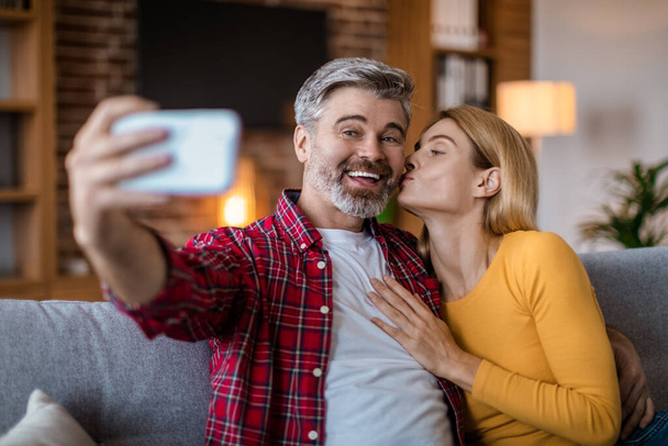 Smiling middle aged caucasian wife kisses husband on cheek, man with beard make selfie on smartphone in room interior. Photo for social networks and blog, video call, love and relationship at home - Foto, immagini