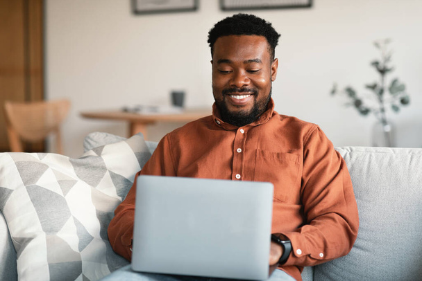 Successful Freelance. Black Male Entrepreneur Using Laptop Typing And Websurfing Working Online Sitting On Couch At Home. Freelancer Guy Browsing Internet On Computer. Technology And Business - 写真・画像