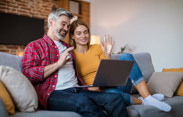 Smiling mature caucasian couple waving hand in laptop, have meeting together, sitting on sofa, in living room interior. New normal at home, video call and social distance with gadget due covid-19 - Fotoğraf, Görsel