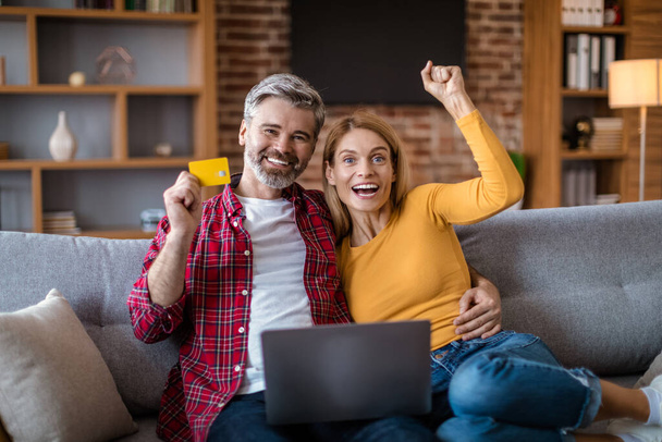 Happy adult caucasian couple with laptop, credit card rejoice to victory, successful purchase, sit on sofa, shopping online in room interior. Sale at home, ad and internet offer, win celebration - Fotó, kép