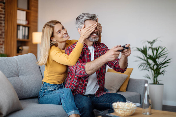 Cheerful mature caucasian lady closes eyes to man with joystick, interferes with online game in free time in living room interior. Fun together at home, entertainment, love and family relationship - Foto, afbeelding