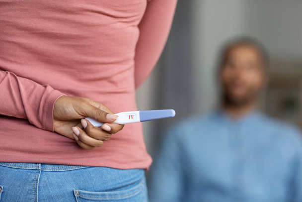Неузнаваемая женщина, скрывающая положительный тест на беременность за спиной, удивляющая своего чёрного мужа дома, афроамериканская пара, готовящаяся к воспитанию, избирательный фокус на женской руке - Фото, изображение