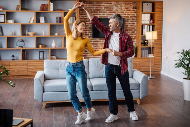 Smiling middle aged caucasian husband and wife dancing, have fun and enjoy free time together in living room interior. Couple celebrating holiday, anniversary at home during covid-19 quarantine - Photo, Image