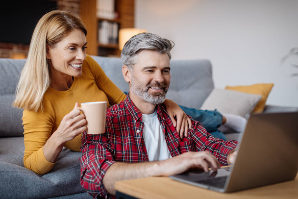 Happy mature caucasian wife and husband looking at laptop, enjoy coffee break in free time in living room interior. Video call, meeting online, modern technology, social network and blog at home - Foto, Imagem