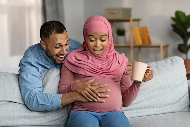 Surprised Pregnant Black Muslim Woman And Her Husband Feeling Baby Kicks Inside Belly, Excited Islamic Spouses Sitting On Couch And Touching Tummy, Emotionally Reacting To Upcoming Parenthood - Photo, image