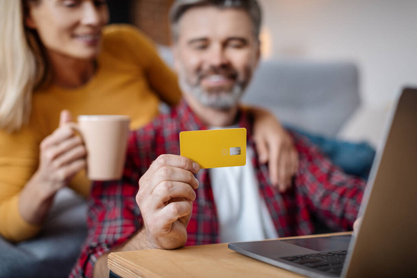 Cheerful mature caucasian lady and man with laptop show credit card and enjoy coffee in living room interior, close up. Break and sale, online shopping, modern technology, purchase order at home - Photo, image