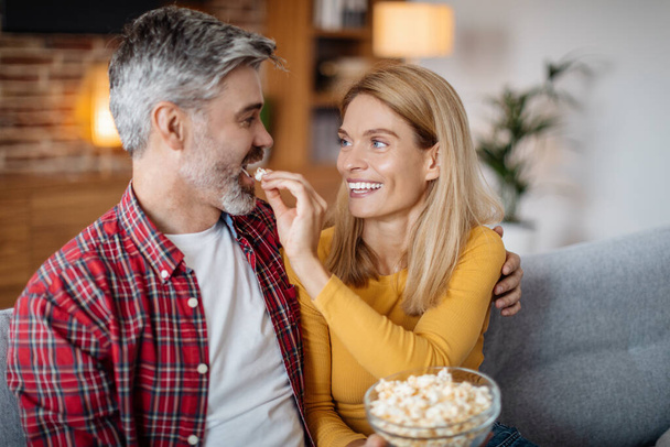 Cheerful middle aged caucasian woman feeding man popcorn, watching tv, enjoy free time in living room interior. Snacks for show and movie evening, entertainment together at home, food and relationship - Foto, afbeelding