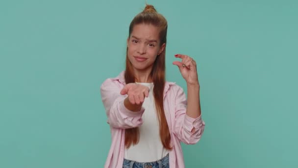 Need some more, please give me. Lovely teenager girl showing a little bit gesture with sceptic smile, showing minimum sign, measuring small size. Young child kid isolated on blue studio background - Imágenes, Vídeo