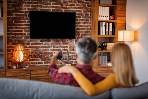Adult caucasian man with remote control and lady relaxing, watching tv with blank screen and enjoy free time in living room interior. Couple and movie night at home together, entertainment with device - Foto, afbeelding