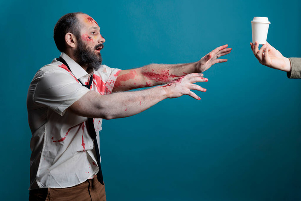 Scary wounded corpse wanting coffee cup in studio over blue background, going after desired drink and acting aggressive. Cruel undead zombie with bloody scars wishing for beverage. - Photo, image