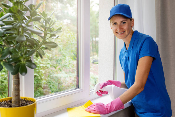 janitorial cleaning services. smiling female worker disinfecting apartment windowsill  - Fotó, kép