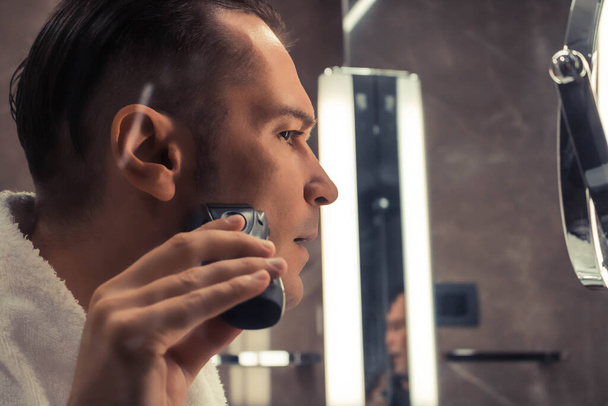 Gros plan de l'homme utilisant un rasoir électrique tout en se rasant dans la salle de bain.  - Photo, image