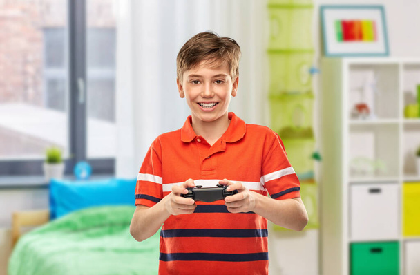 gaming, leisure and people concept - portrait of happy smiling boy in red polo t-shirt with gamepad playing video game over home room background - Photo, Image