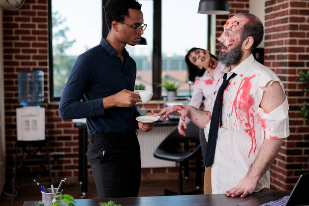 Evil zombie talking to businessman in office, bloodthirsty brain eating monster having conversation in business workplace. Walking dead corpse with bloody wounds and sinister scars. - 写真・画像