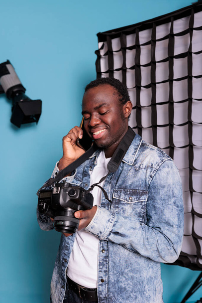 Confident photography enthusiast having a phone call in professional production studio on blue background. Happy photographer talking on smartphone while having a photo shooting. - Foto, Imagem