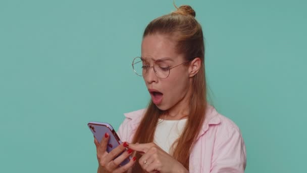 Sad teenager student girl in glasses use smartphone typing browsing, loses becoming surprised sudden lottery results, bad fortune, loss. Young child kid isolated on blue studio background indoors - Video