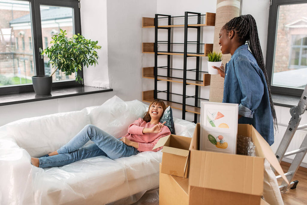 moving, people and real estate concept - happy smiling woman unpacking box and her friend resting on sofa at new home - Φωτογραφία, εικόνα