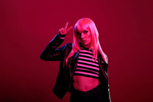 Portrait of amazing female model doing peace sign over dark pink light in studio. Cool casual woman wearing pink hair wig, natural leather jacket and stylish sunglasses, showing victory gesture. - Foto, immagini