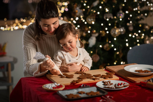 family, cooking and winter holidays concept - happy mother and baby daughter decorating gingerbread cookies at home on christmas - Fotografie, Obrázek