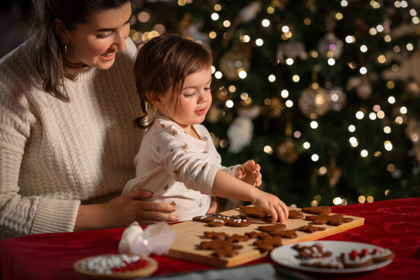 family, cooking and winter holidays concept - happy mother and baby daughter with icing in baking bag decorating gingerbread cookies at home on christmas - Foto, Bild