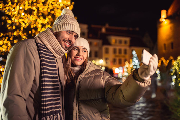 winter holidays and people concept - happy smiling couple taking selfie with smartphone over christmas tree lights in evening city - Foto, afbeelding