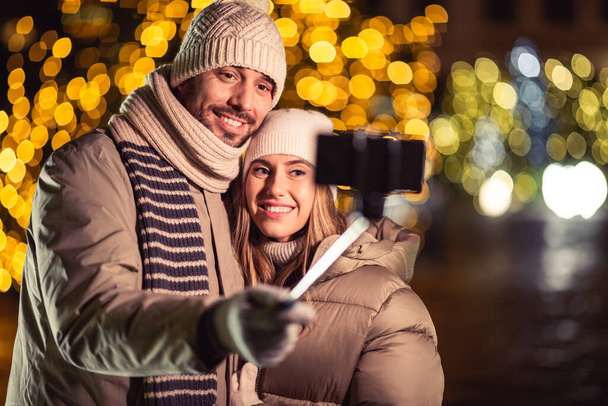 winter holidays and people concept - happy smiling couple taking picture with smartphone on selfie stick and hugging over christmas lights in evening - Photo, Image