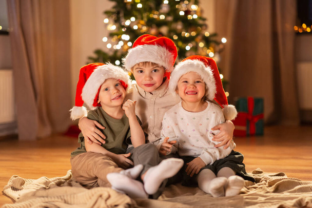 christmas, winter holidays and childhood concept - happy little girl and boys in santa hats hugging on floor at home - 写真・画像