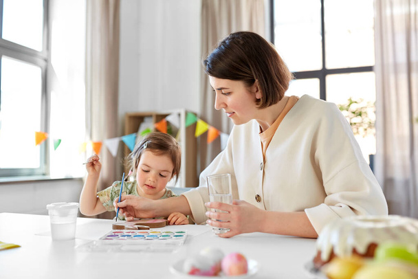 holidays, family and people concept - happy mother and little baby daughter coloring easter bunny with watercolors at home - 写真・画像