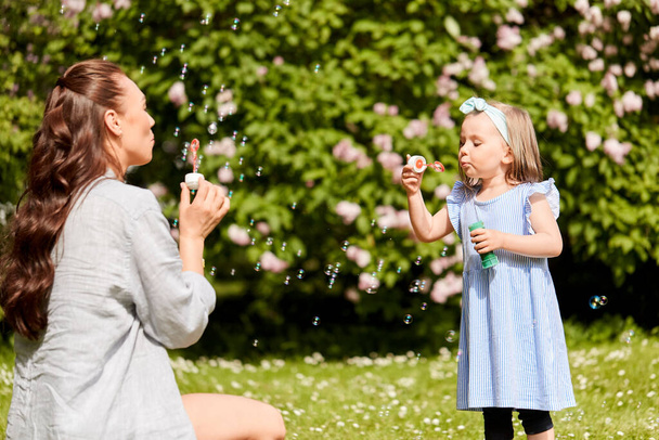 family, motherhood and people concept - happy mother with little daughter blowing soap bubbles at summer park or garden - Foto, afbeelding