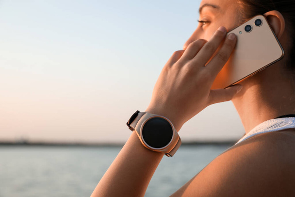 Woman with fitness tracker talking on phone near river, closeup. Space for text - Foto, immagini