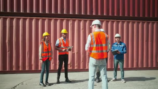 Группа многорасовых рабочих в униформе безопасности, стоящих в ряд и обсуждающих с кавказским менеджером на логистическом причале со стопками контейнеров, судоходных грузов и грузовых перевозок. - Кадры, видео