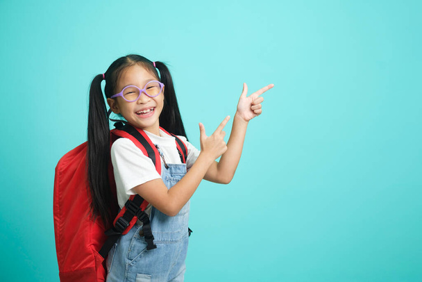 Χαμογελαστό κοριτσάκι με γυαλιά που δείχνει το δάχτυλο προς τα πάνω πηγαίνοντας σχολείο, σχολική ιδέα. στέκεται σε μπλε φόντο. - Φωτογραφία, εικόνα