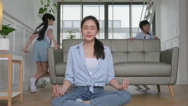 Young Asian Thai mother sits on the living room floor, meditates, and practices yoga for health and wellness, the children play, chaotic and naughty, happy domestic home lifestyle on family weekend. - Video