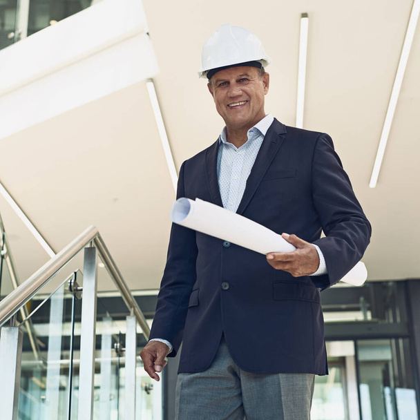 Without him there will be no development. Portrait of a cheerful professional male architect looking at the camera while holding blueprints inside a building - Fotoğraf, Görsel