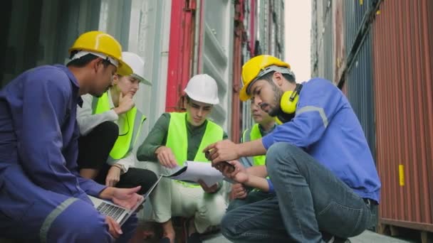 Group of multiracial workers people in safety uniforms and hardhats sit and discuss in stacks of logistics containers with male White manager, trading shipping goods for cargo transportation industry. - Imágenes, Vídeo