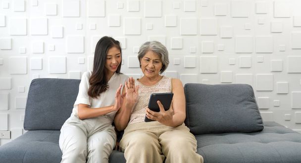 Молода азіатка і її бабуся махають рукою через віддалений зв'язок через планшетний комп'ютер у вітальні. Щаслива сім'я, що живе разом
 - Фото, зображення
