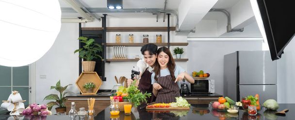 Asian couple spend time together in the kitchen. Young woman in apron cooking salad dish while his boyfriend recording vlog video for social blogger. Photography studio lights set up in the kitchen. - Foto, Imagem