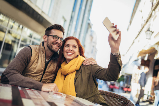 Ευτυχισμένο νεαρό ζευγάρι που χρησιμοποιούν ένα τηλέφωνο μαζί σε μια καφετέρια, κάνουν selfie φωτογραφία. - Φωτογραφία, εικόνα