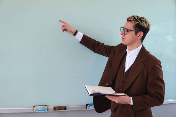 A teacher pointing at a blackboard. University professor with a black book in his hand. Master dressed in an elegant brown suit. Elegant man in a blackboard explaining a topic. High quality photo - Foto, afbeelding