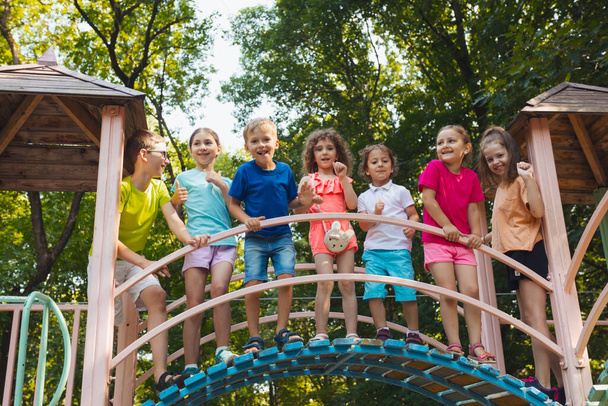 The elementary school children stand on a bridge in the playground and hold on to the railing. The kids dressed in colorful clothes are posing for the camera - Photo, Image