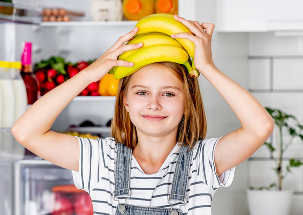 Preteen girl with bananas vitamin healthy food at kitchen. Pretty child kid enjoying yellow fruits at home - Photo, Image