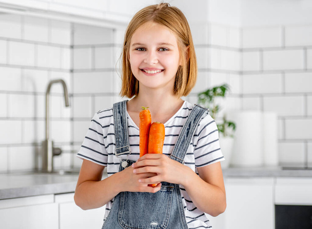 Preteen girl holding carrots at kitchen and smiling. Pretty child with orange vegetables food at home - Fotoğraf, Görsel