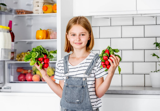Preteen girl with red radish at kitchen posing and smiling. Pretty child with fresh vitamin vegetables at home - Photo, Image