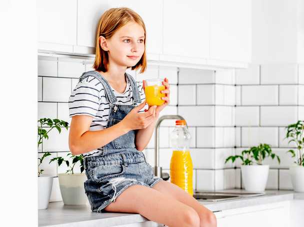 Preteen girl with orange juice bottle and glass sitting at kitchen table. Pretty child kid with vitamin fruit drink at home - Foto, Imagen