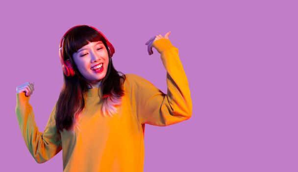 Young asian woman in yellow sweatshirt wearing red headphones listen to music and dancing on the pink purple color background. - Foto, Imagem