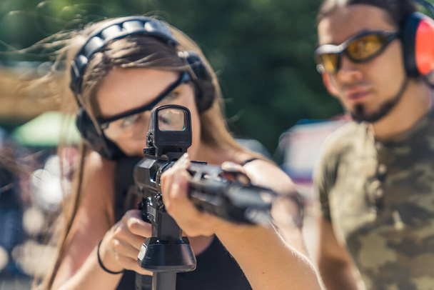 Strong long-haired caucasian girl pursuing her hobby by shooting from a black rifle with collimator. Male instructor in the background. Closeup portrait. Blurred foreground. . High quality photo - 写真・画像