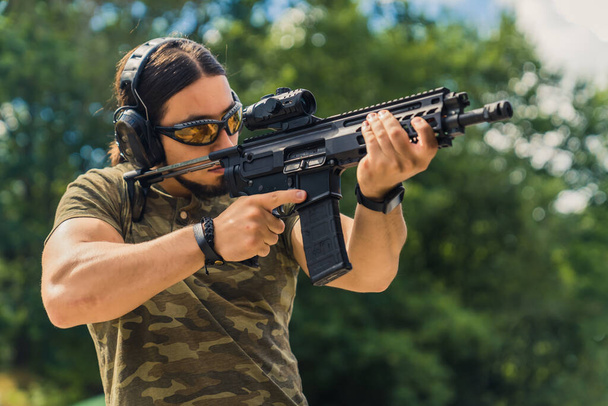 Handsome male instructor at shooting range. Medium outdoor shot of a muscular caucasian focused man in protective eyeglasses and headphones aiming at target with a rifle looking through the loupe - Foto, immagini
