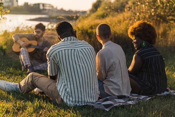 Rear view of a multicultural group of friends enjoying pleasurable sunset time in nature near the river while sitting on the grass, talking and laughing. A young bearded man playing the guitar. High - Fotografie, Obrázek