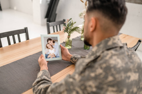 Uniformed man sitting alone at the kitchen table with the family photo in his hands - Foto, Bild